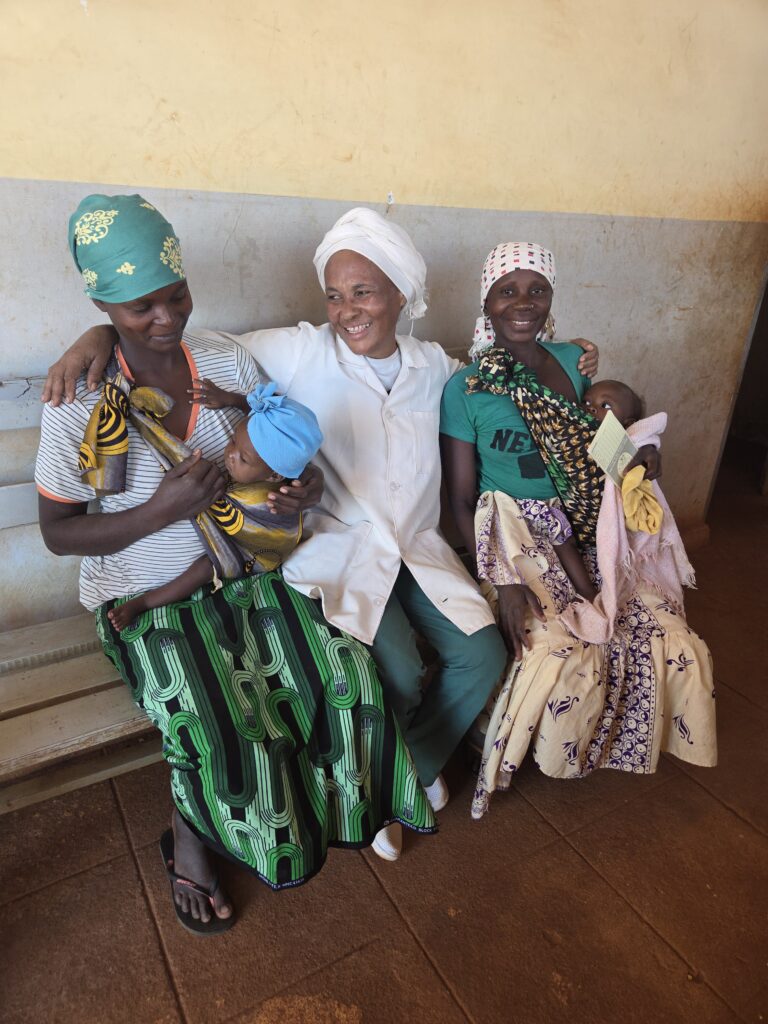 Smiling midwife in Mozambique sits with two mothers holding their babies at a rural health clinic supported by a Canadian charity.