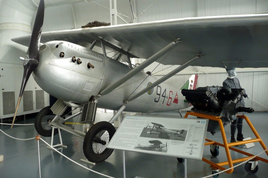 Vintage silver biplane with propeller on display in an aviation museum alongside an engine and historical exhibit signage.