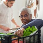 Older Black man receiving fresh vegetables from a community food program, with a younger volunteer assisting in the background&mdash;representing food security and care provided by charities in B.C