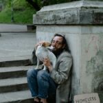 A man experiencing homelessness sits on stone steps hugging his small dog, beside a cardboard sign that reads &ldquo;Homeless and Hungry.&rdquo;
