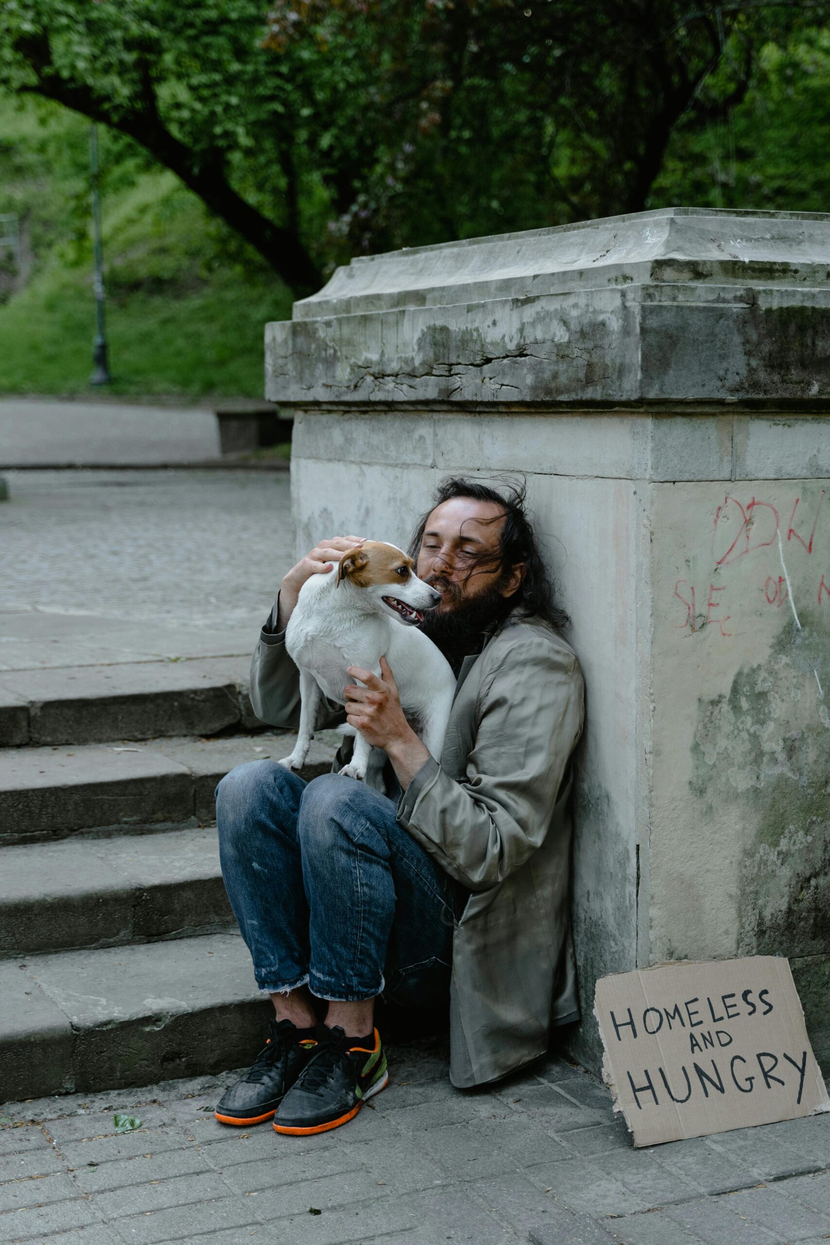 A man experiencing homelessness sits on stone steps hugging his small dog, beside a cardboard sign that reads &ldquo;Homeless and Hungry.&rdquo;
