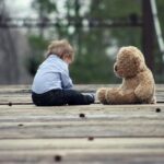A young child sits on a wooden bridge facing a teddy bear, creating a quiet and emotional scene.