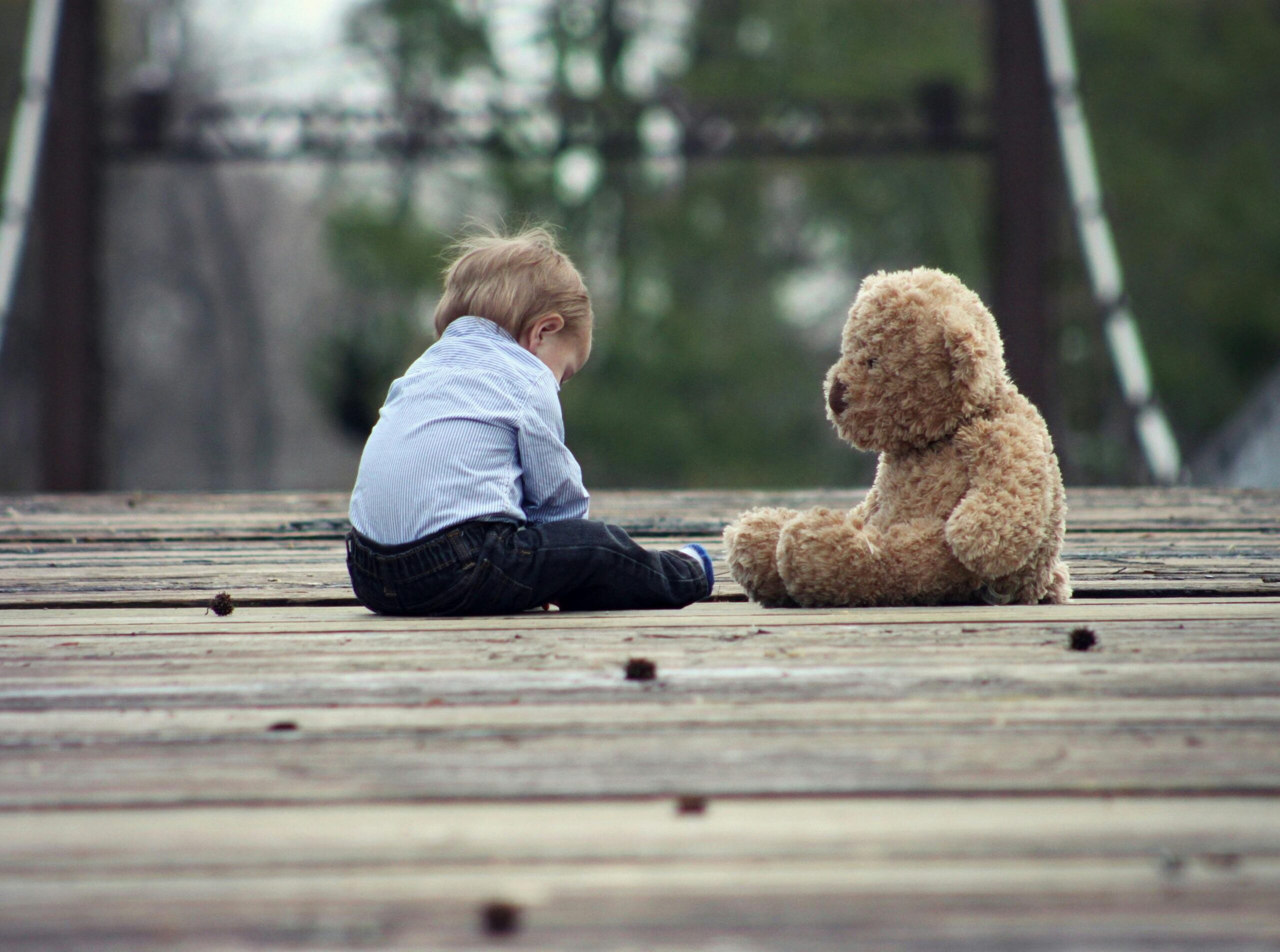 A young child sits on a wooden bridge facing a teddy bear, creating a quiet and emotional scene.