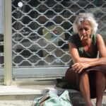 Older woman sitting on a sidewalk with her belongings, illustrating homelessness and housing instability after hospital discharge.